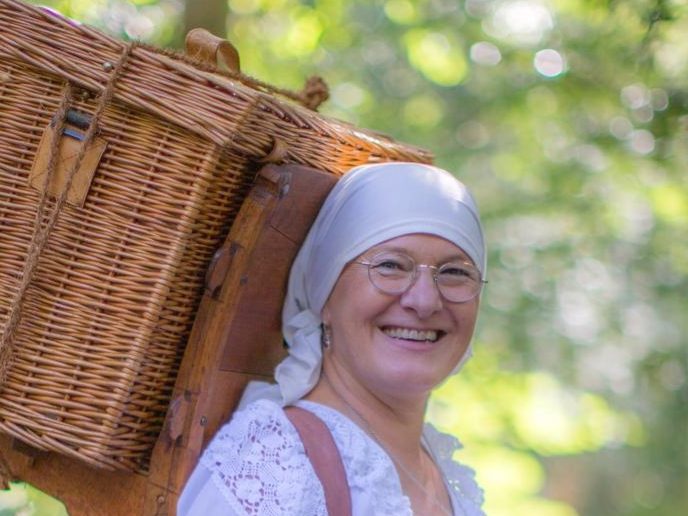 Bucklapoteca Maike Frau mit einem Korb auf dem Rücken, lächelt freundlich in einer grünen Umgebung.