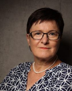Porträt einer Frau mit kurzen, dunklen Haaren, Brille und heller Bluse, mit ernstem Blick.
